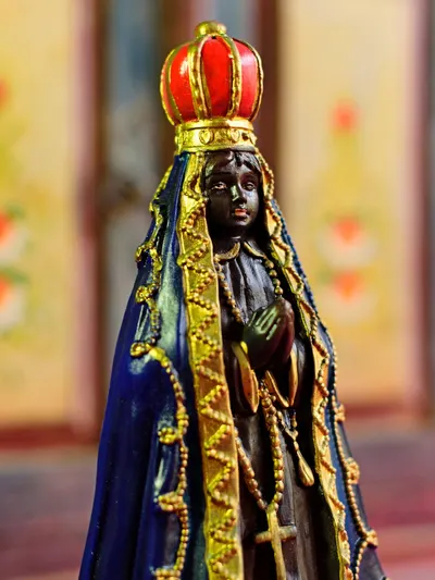 Foto da imagem de Nossa Senhora Aparecida, escura, de manto azul com orla dourada e coroa vermelha, mãos em prece.