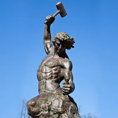 Escultura de bronze de homem musculoso que se esculpe da própria pedra, martelo erguido, contra o céu azul.